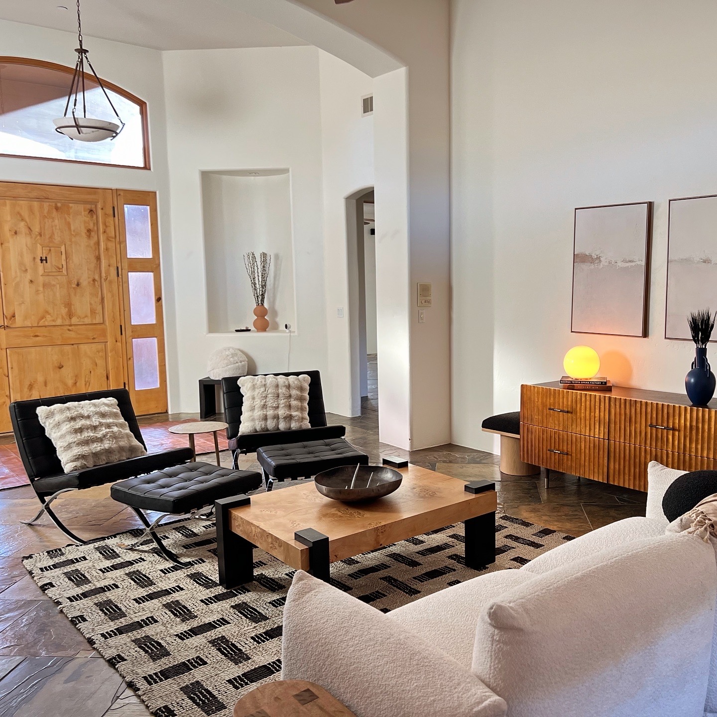 Desert modern living room staged by Desert Varnish Design in Sedona featuring Barcelona chairs with black leather, burl wood coffee table, fluted wood credenza, bouclé sofa, geometric woven rug, slate tile floors, knotty alder front door with arched transom window, and nicho wall detail in a southwestern adobe-style home